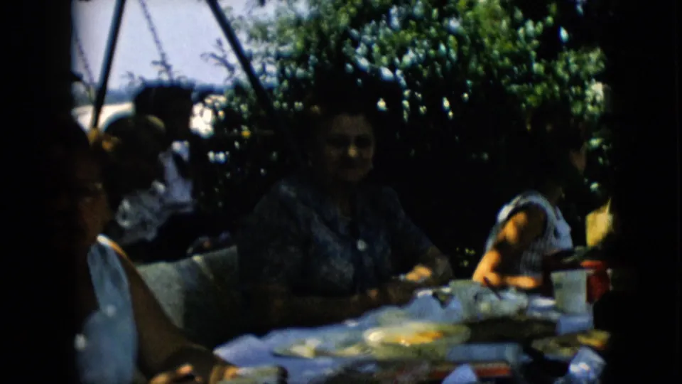 1957 Pennsylvania frame focused on a woman in a patterned blouse seated at the outdoor picnic table, with tableware, food containers, greenery, and partial family figures around her.