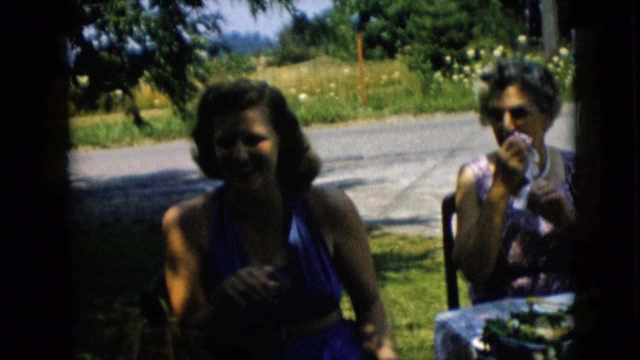 In 1957 Pennsylvania home-movie footage, two adult women sit at a small shaded picnic table near a quiet road, one in a blue halter dress and the other in a patterned dress.