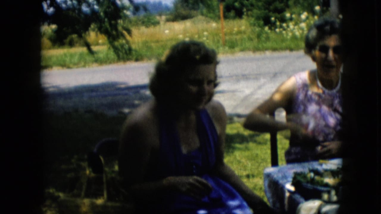 Two women sit at a picnic table with grass, trees, and road behind them.