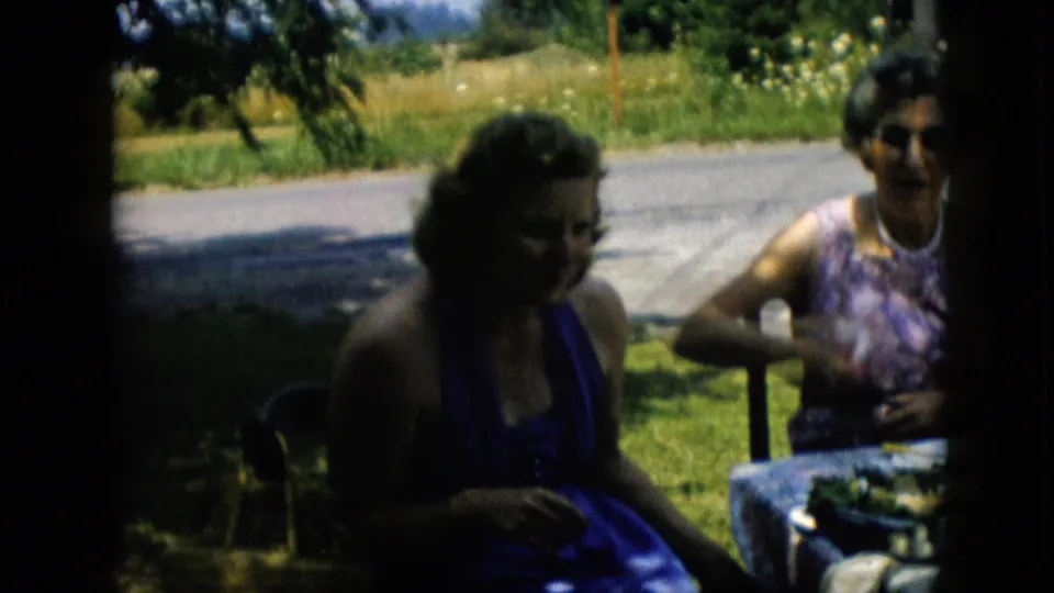 Two adult women remain seated outdoors near a roadside lawn, with the woman in the patterned dress raising food or a hand near her mouth.