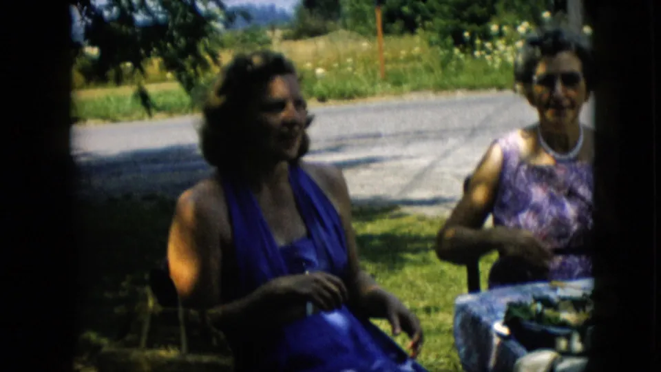 A candid 1957 Pennsylvania frame shows two adult women smiling at a shaded picnic table, with road pavement and bright grass in the background.