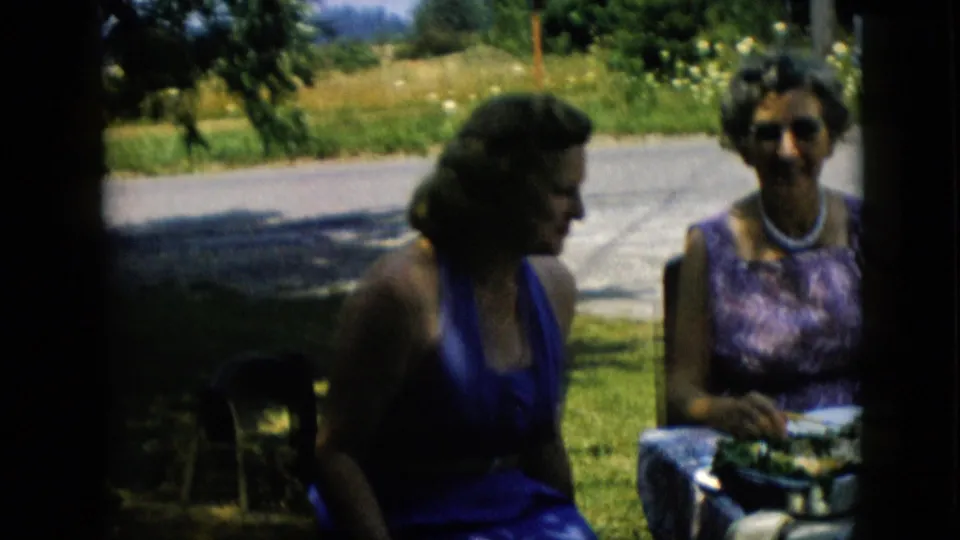 The woman in the blue dress turns in profile while the woman in the patterned dress faces forward at a shaded outdoor table in the supplied 1957 Pennsylvania home-movie clip.