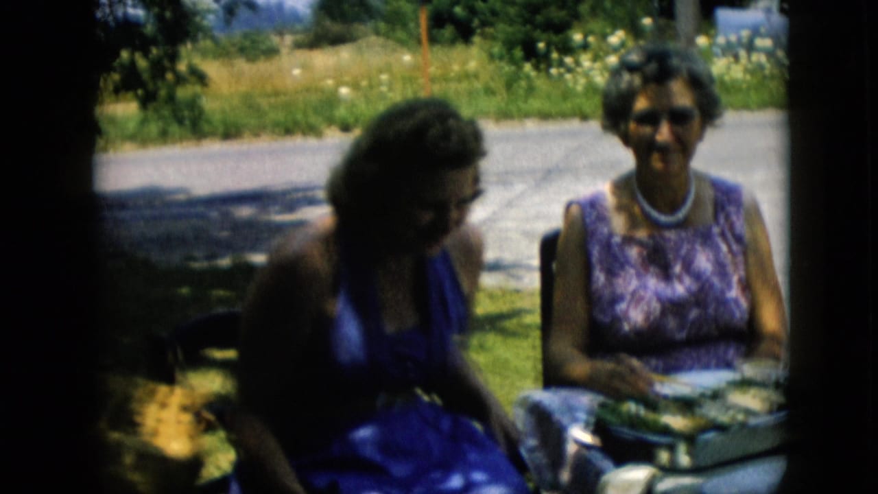 Two women sit outdoors at a table with serving dishes and plates in 1957 Pennsylvania home-movie footage, with grass and a road behind them.