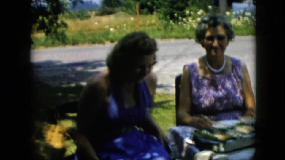 Two women sit outdoors at a table with serving dishes and plates in 1957 Pennsylvania home-movie footage, with grass and a road behind them.