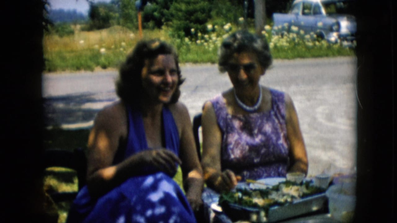 Two women smiling at an outdoor table with food and parked cars behind them.