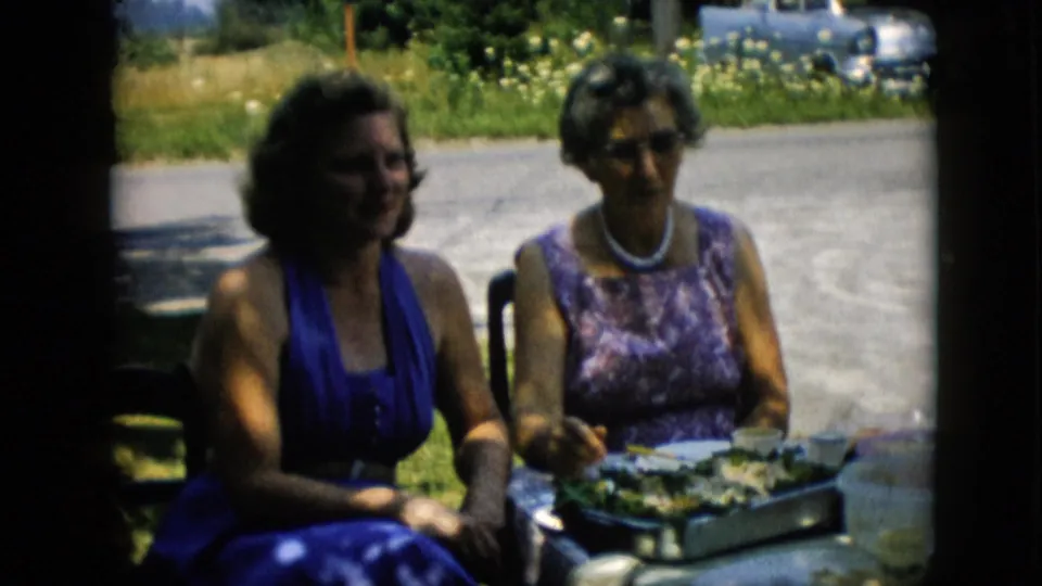 Two women sit side by side at a sunny outdoor brunch table in Pennsylvania home-movie footage, with serving dishes, grass, and roadside background visible.