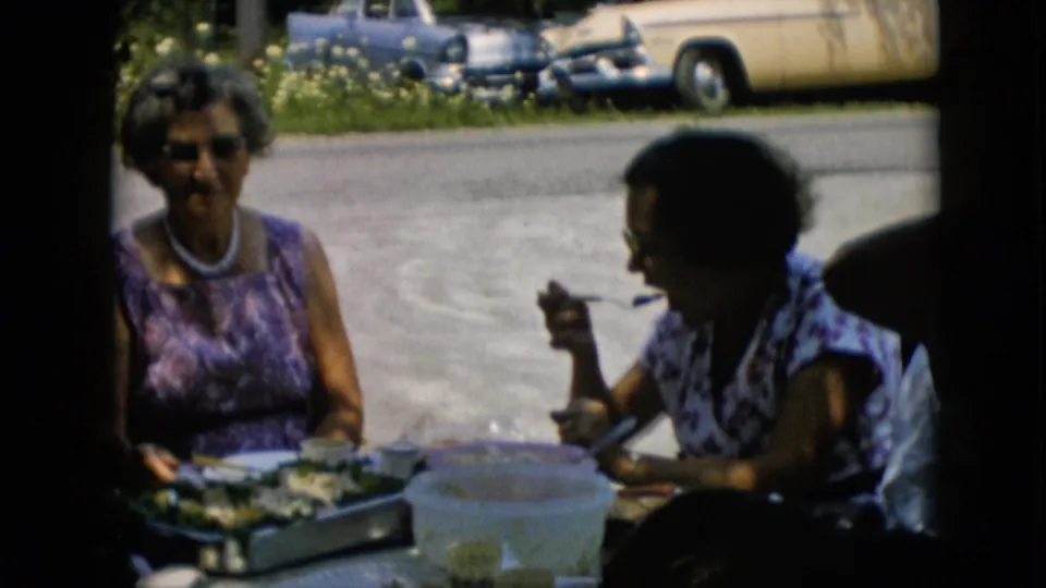 An older woman in a purple patterned dress faces the camera while another woman eats at the outdoor table, with a pitcher and parked cars behind them.