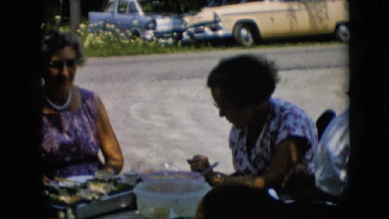 In 1957 Pennsylvania home-movie footage, two adult women sit at a shaded outdoor table with plates and containers as midcentury cars are parked in the sun behind them.