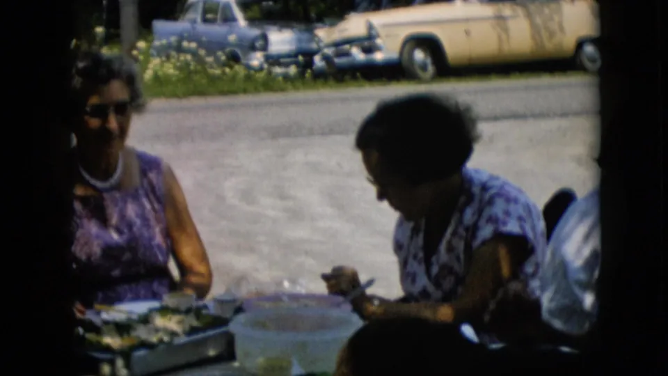 In 1957 Pennsylvania home-movie footage, two adult women sit at a shaded outdoor table with plates and containers as midcentury cars are parked in the sun behind them.