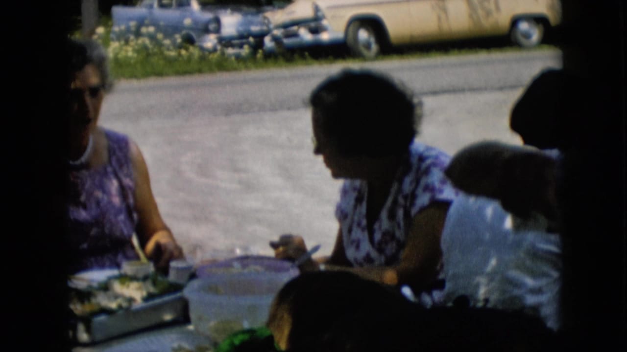 Women at a shaded table eat and talk with a yellow car visible across the road.