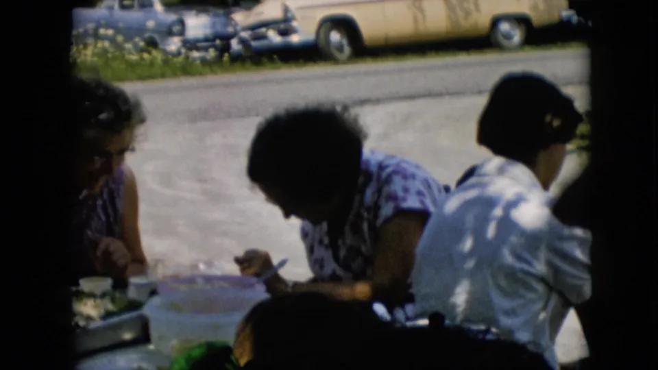 The home-movie frame shows adult diners gathered closely around the outdoor table, with a woman in a patterned top eating and midcentury cars parked beyond the gravel drive.