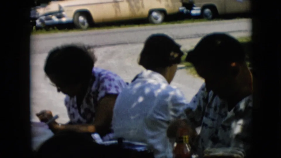 Several adult figures sit close together at the shaded meal table, including a woman in a white shirt and another in a patterned blouse, with a yellow car visible across the driveway.