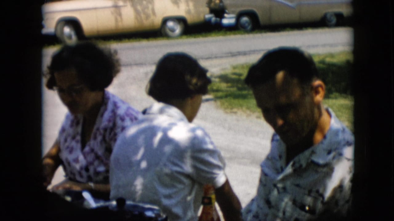 In this Pennsylvania 1957 home-movie frame, three adults sit outdoors in shade near parked 1950s sedans, with the man in a patterned shirt partly turned toward the camera.