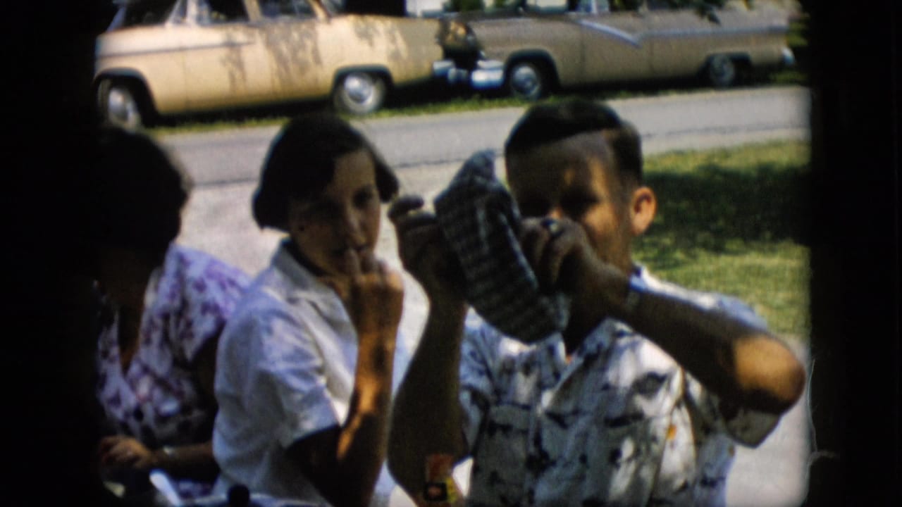 A seated man lifts a checkered cap near his head while two women sit beside him outdoors.
