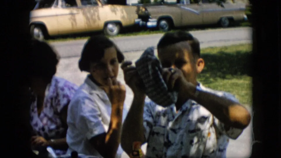 In this Pennsylvania 1957 frame, a man in a patterned shirt raises a blue and white checkered cabbie-style cap while a woman in a white blouse smiles nearby and another woman sits partly in shadow.