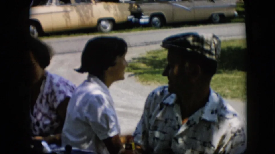In this Pennsylvania 1957 home-movie frame, the man now wears the checkered cap while seated beside two women, with a residential street and parked cars in the background.