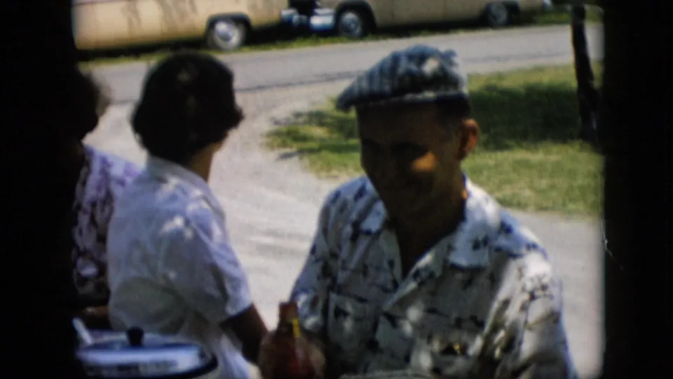 In this Pennsylvania 1957 frame, the man in the checkered cap sits at the edge of the group while two women turn away slightly, with lawn, driveway, and parked sedans visible behind them.