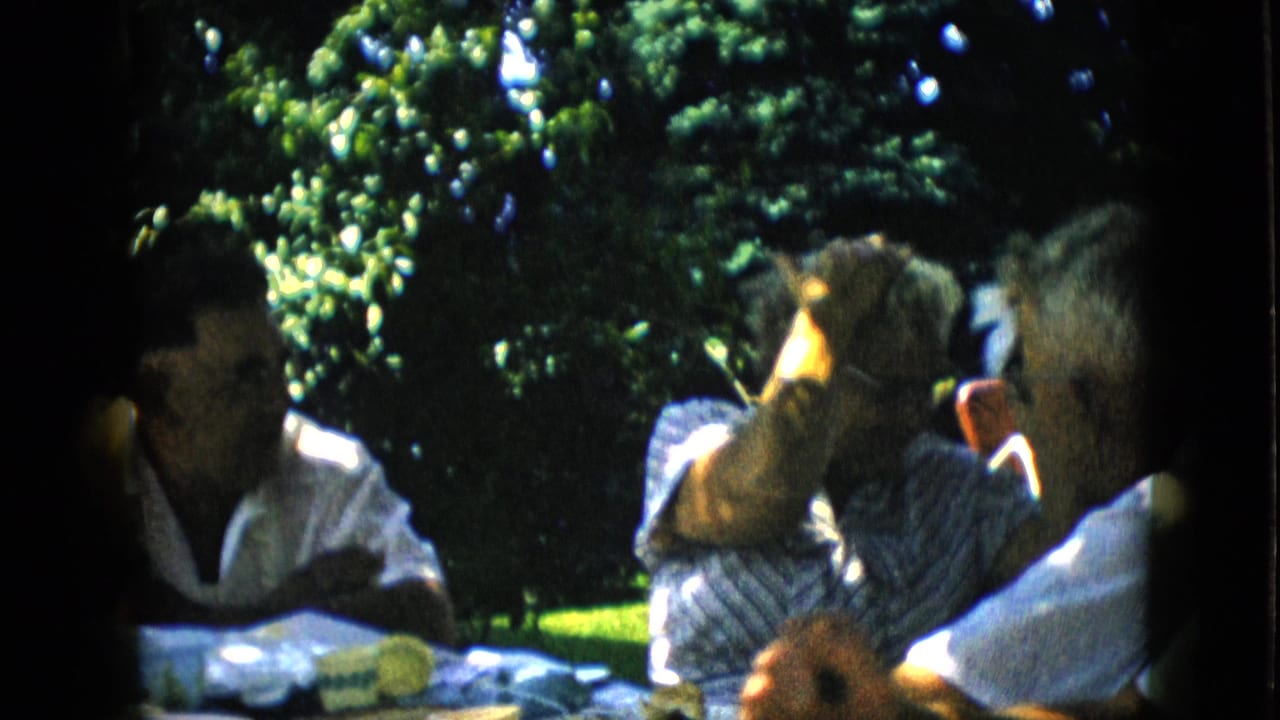 1957 Pennsylvania 8mm home-movie frame showing adults seated around an outdoor table in dappled garden light, with leafy trees behind them.