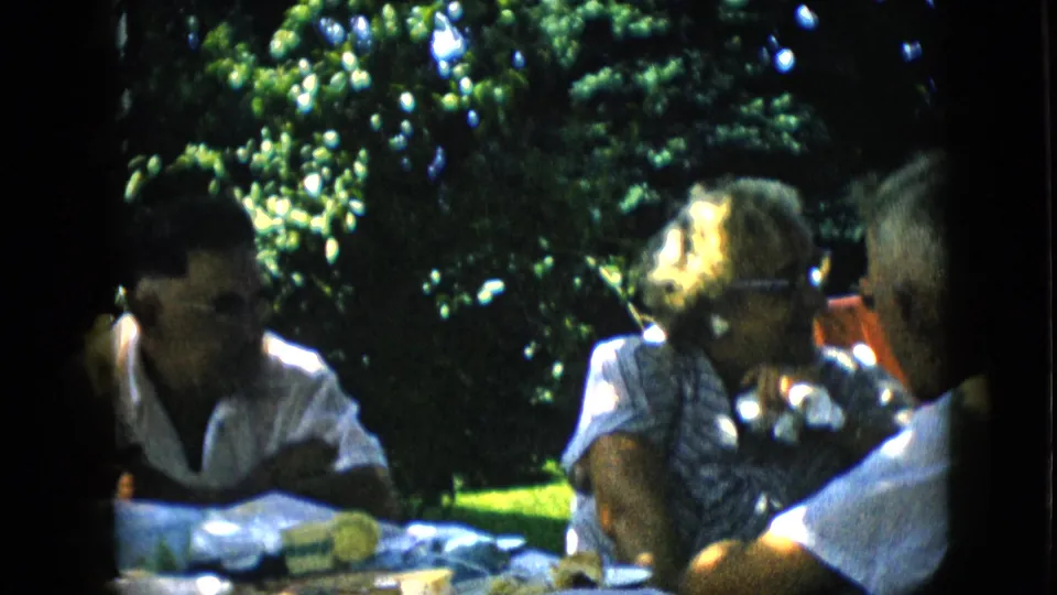 1957 Pennsylvania home-movie still of a woman in glasses and a patterned blouse turning during conversation with men at a shaded outdoor table.