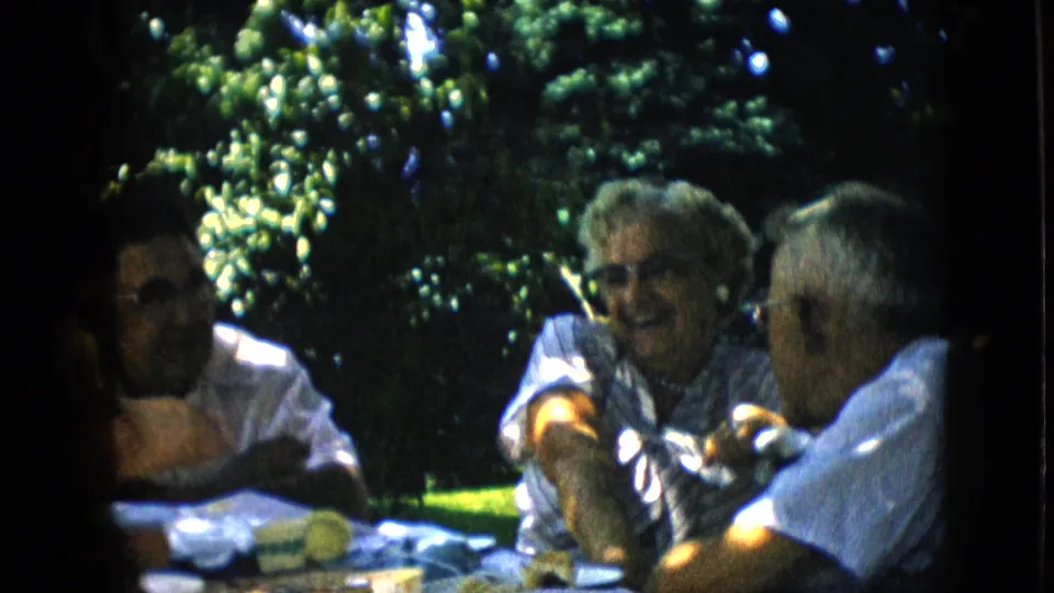 1957 Pennsylvania 8mm frame showing adults gathered at a table outdoors, with the woman smiling between two men and leafy trees in the background.