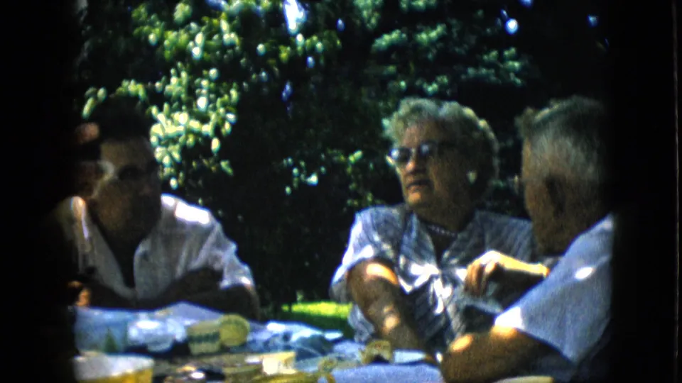 1957 Pennsylvania home-movie still of a woman wearing sunglasses seated at a garden table with two men, framed by dark green trees and bright sunlight.
