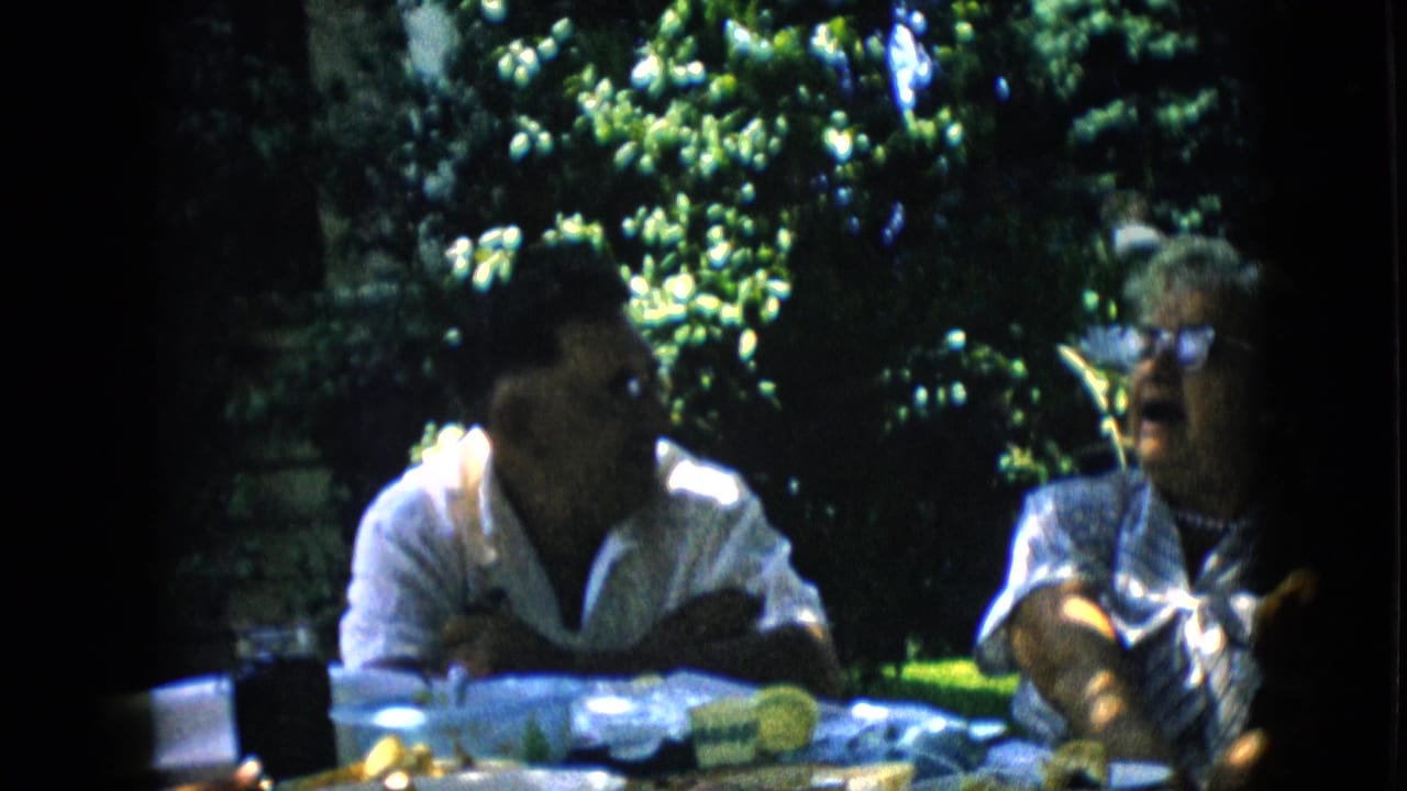 In 1957 Pennsylvania home-movie footage, two adults sit at a shaded backyard table with food and cups while talking beneath leafy trees.