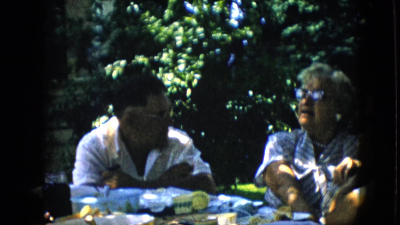 Two adults seated at a table outdoors, with one person gesturing while they talk.