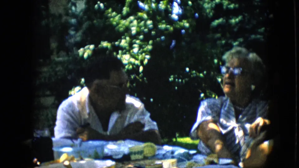 Two adults sit at a backyard table in shaded daylight, one gesturing during conversation as food and cups rest on the table.