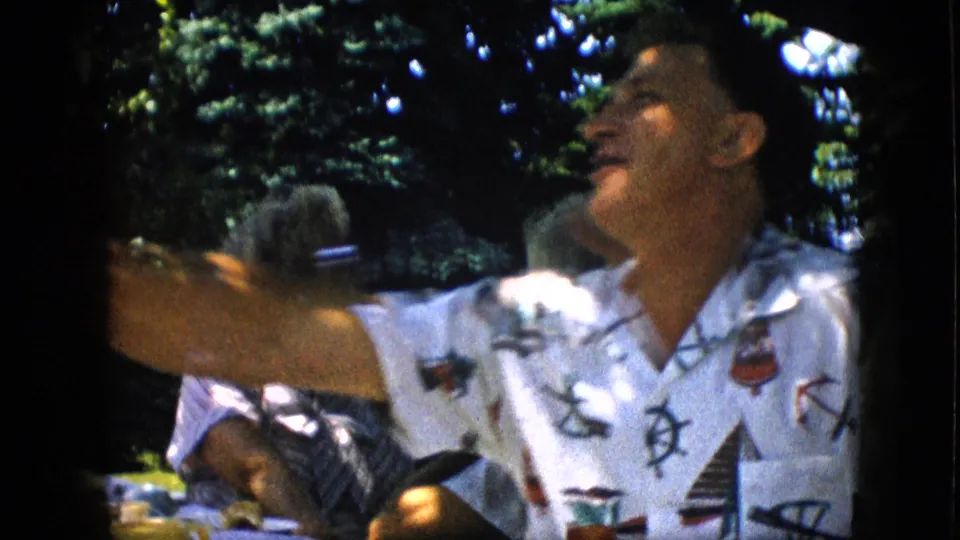 A man in a white nautical-patterned shirt raises his arm in a lively backyard party moment, with other seated adults and trees behind him.