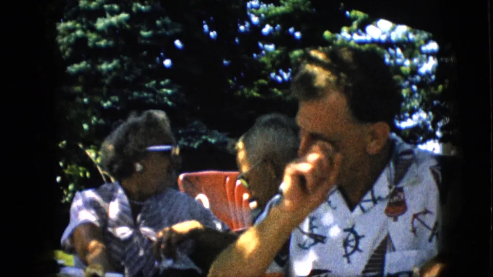 A man in a white nautical-patterned shirt sits beside a red outdoor chair while other adults continue the shaded backyard table gathering.