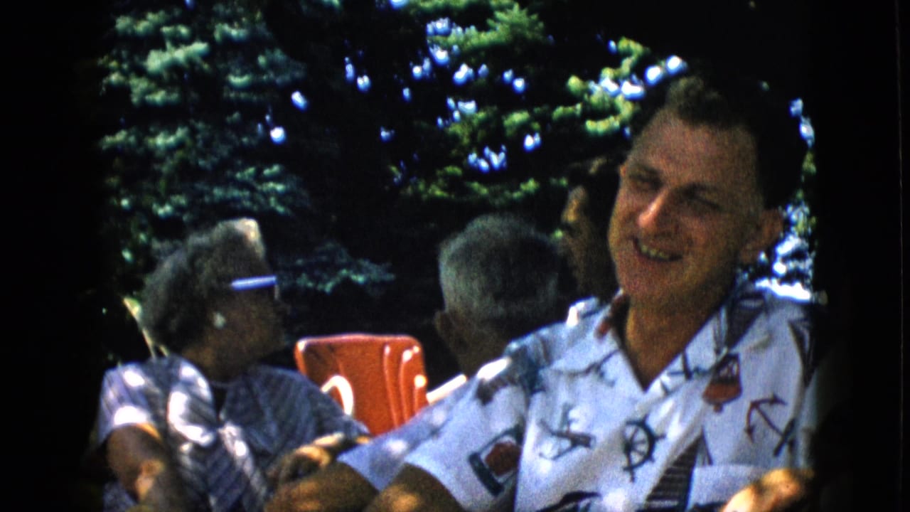In supported 1957 Pennsylvania home-movie footage, a man in a white nautical-patterned shirt laughs at an outdoor gathering while an older woman in sunglasses and a red chair appear behind him.