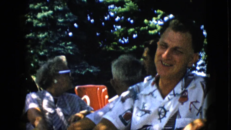 In supported 1957 Pennsylvania home-movie footage, a man in a white nautical-patterned shirt laughs at an outdoor gathering while an older woman in sunglasses and a red chair appear behind him.