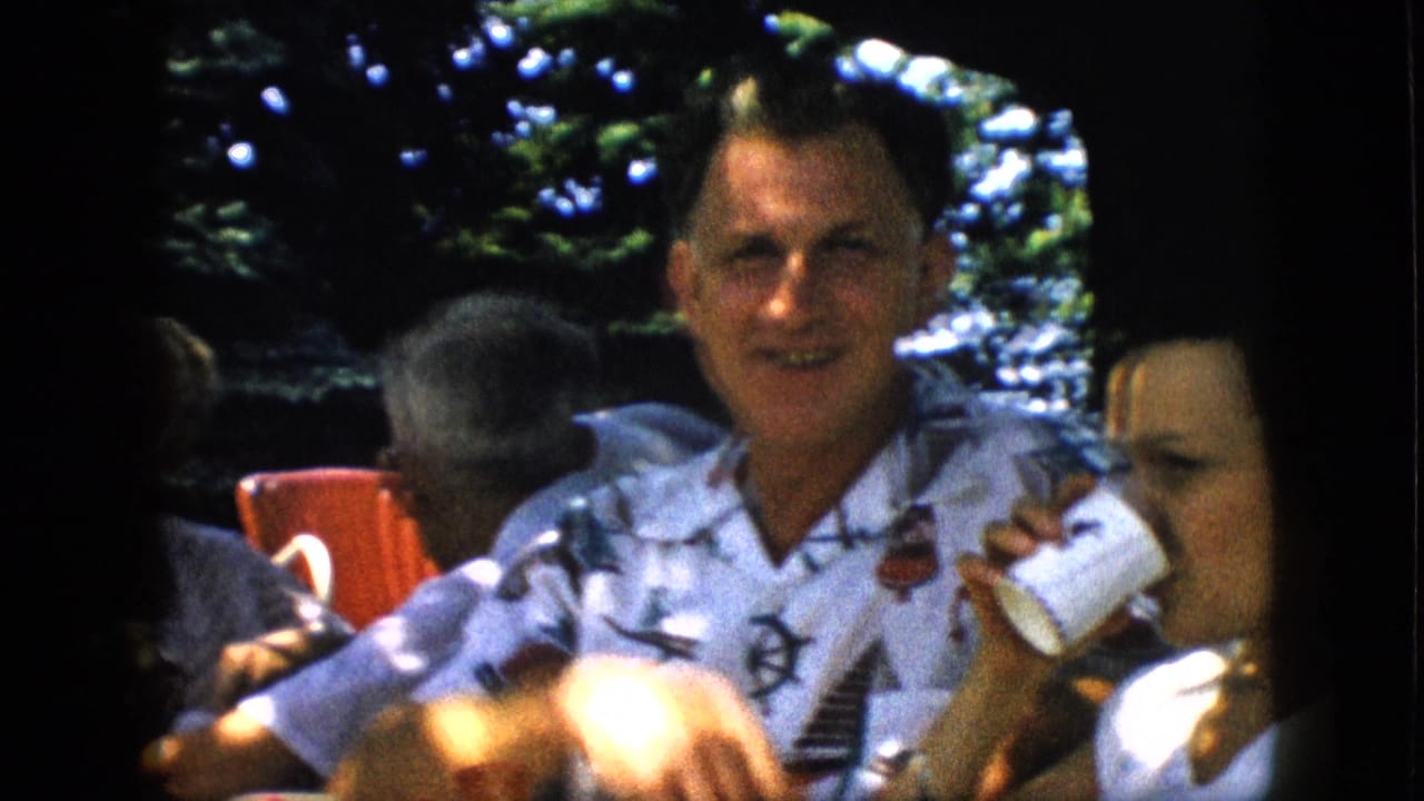 A smiling man in a white patterned shirt gestures beside a woman in a white top at a 1957 Pennsylvania outdoor family gathering, with red chairs and trees in the background.