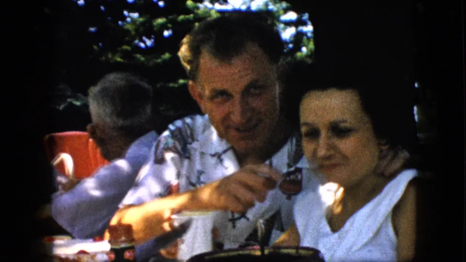 The man leans forward and points while a woman in a white sleeveless top sits beside him, both framed in soft 8mm color at a 1957 Pennsylvania outdoor gathering.