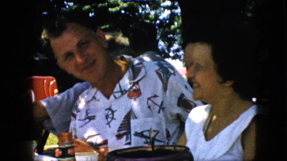 A man in a white patterned shirt and a woman in a white top smile together at an outdoor table in supported 1957 Pennsylvania home-movie footage, with cups and shaded greenery visible.