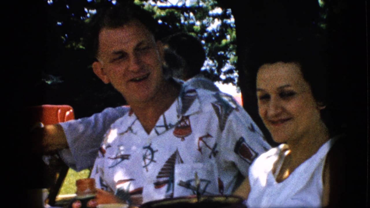 In 1957 home-movie footage from the United States, a man in a white nautical-pattern shirt holds a red cup beside a woman in a white top during an outdoor family picnic.