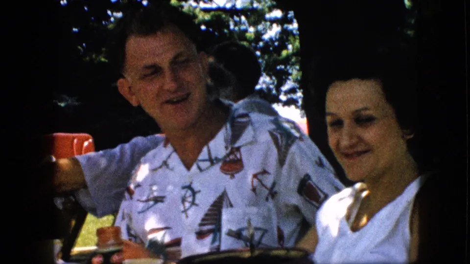 In 1957 home-movie footage from the United States, a man in a white nautical-pattern shirt holds a red cup beside a woman in a white top during an outdoor family picnic.