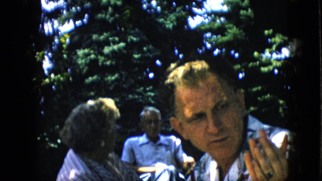 Blond man looks toward the camera while other adults sit behind him under trees.