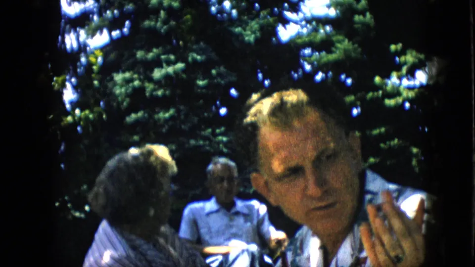 A blond man in a light patterned shirt faces the camera at a 1957 outdoor family picnic, with other seated adults visible in soft focus beneath leafy trees.