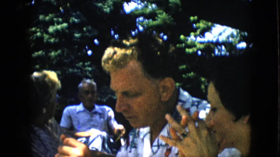 A man appears in profile during the 1957 family reunion picnic, seated near other adults in dappled sunlight with leafy trees and soft background figures behind him.
