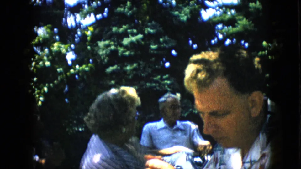Adults remain seated under trees at the 1957 family picnic as a foreground man looks down, with background relatives softly visible in the shaded outdoor setting.