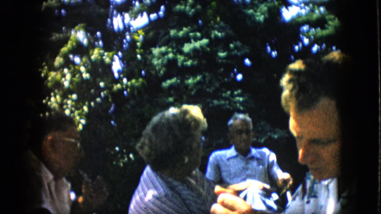 In metadata-supported 1957 Pennsylvania home-movie footage, several adults sit outdoors in a leafy backyard while conversing in dappled sunlight.