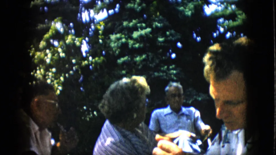 In metadata-supported 1957 Pennsylvania home-movie footage, several adults sit outdoors in a leafy backyard while conversing in dappled sunlight.