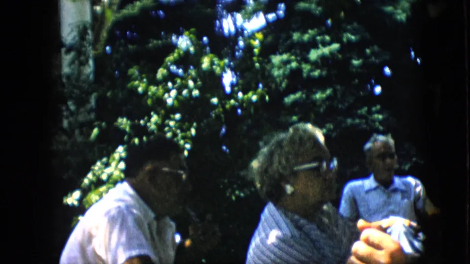 The frame shows adults in a shaded Pennsylvania backyard, with an older woman in profile, a man with glasses nearby, and another man seated behind them.
