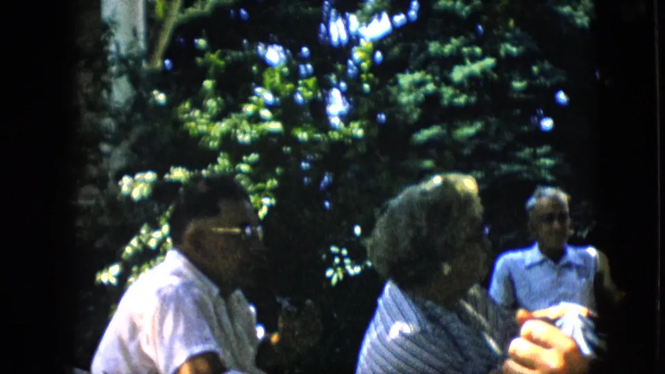 A handheld 8mm frame shows seated adults in profile under tall backyard trees, with a man in a light shirt visible in the background.