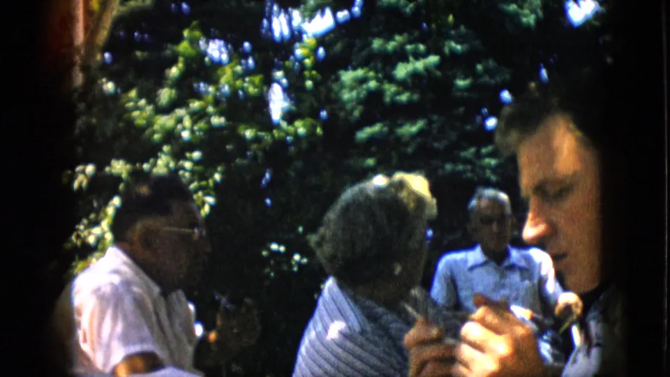 In the final frame, a man appears close to the camera while other adults remain seated in the shaded backyard, continuing the informal outdoor gathering.