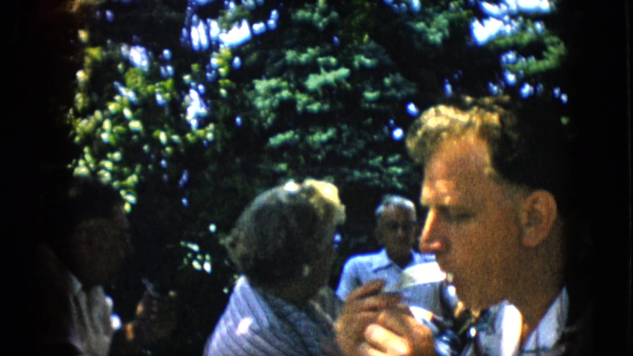 In metadata-supported 1957 Pennsylvania home-movie footage, adults sit outdoors beneath trees while a man in a patterned shirt eats in close view.
