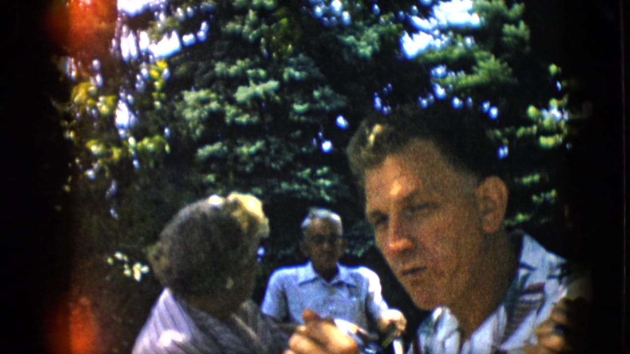 Man in patterned shirt faces the camera while adults sit behind him outdoors.