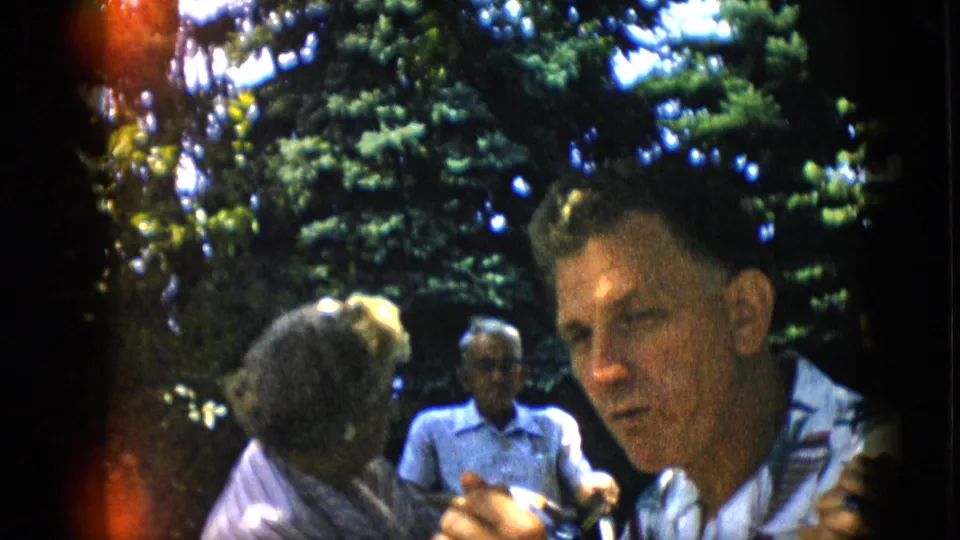 A man in a patterned shirt turns toward the camera at an outdoor Pennsylvania gathering, with seated adults and trees visible behind him.