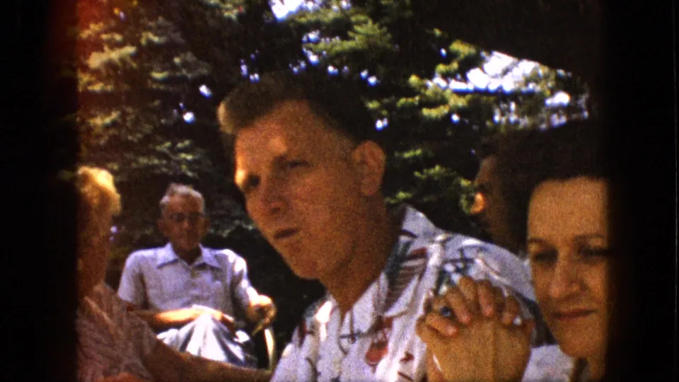 The home-movie frame shows a man in a patterned shirt seated close to a dark-haired woman, with other adults and trees in the background.
